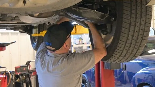 A mechanic in San Antonio works under a raised car, inspecting or repairing near the rear wheel in a garage—capturing a slice of everyday history beneath the blue moons of Texas.