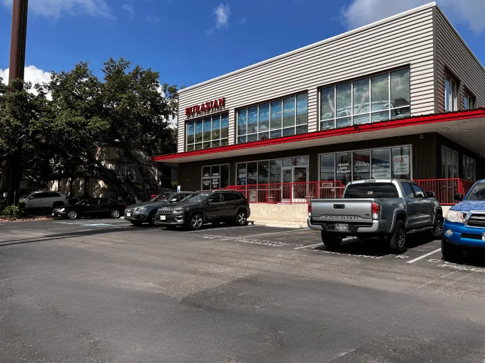 A two-story building with large windows and a red awning labeled “PHARMACY” stands beside the Eurasian Auto Repair shop—a San Antonio auto repair shop—where several cars are parked in the adjacent lot under a partly cloudy sky.