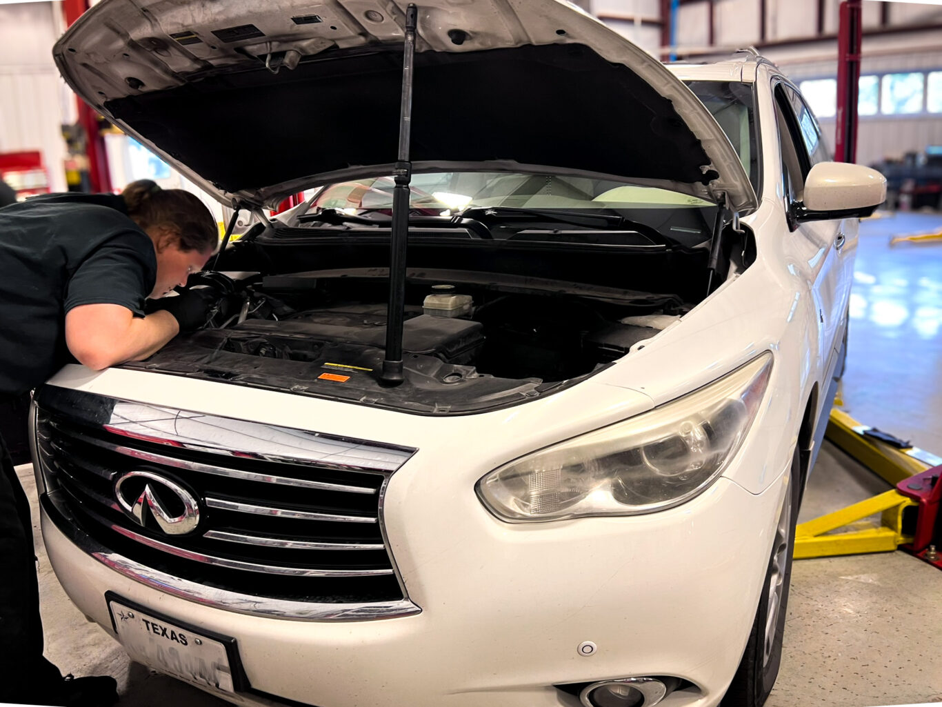 A mechanic inspects the engine of a white Infiniti SUV inside a San Antonio auto repair shop.