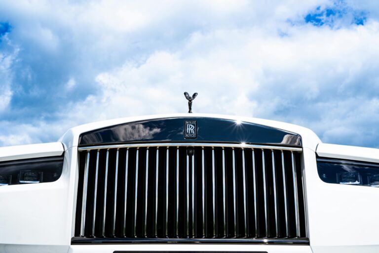 Close-up of the front grille and emblem of a white Rolls-Royce, highlighting its elegance among luxury SUVs, with a partly cloudy sky in the background. In the background, subtly incorporate elements that suggest it's parked at a San Antonio auto repair shop