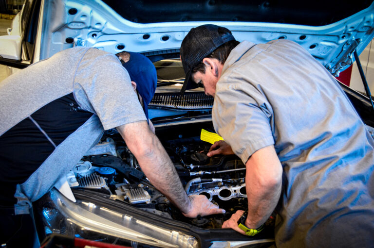At a San Antonio auto repair shop, two men wearing caps and work shirts lean over the open hood of a car, inspecting and working on the engine components.