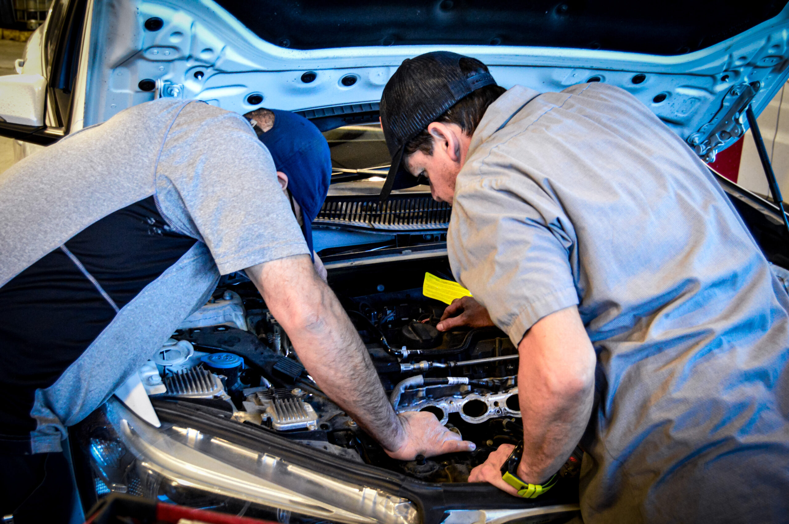 At a San Antonio auto repair shop, two men wearing caps and work shirts lean over the open hood of a car, inspecting and working on the engine components.
